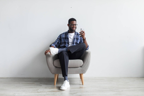 Chico negro feliz sentado en sillón con teléfono inteligente, mensajes de texto a amigo, enviar mensaje, navegar por la web contra la pared blanca - Foto, imagen