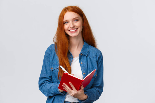 Waist-up portrait lovely redhead teenage girl taking notes during favorite college class, holding pen and red notebook, smiling carefree camera, writing something, make to-do list - Foto, Bild