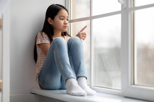 Unhappy school-aged korean girl sittng on windowsill at home - Photo, Image