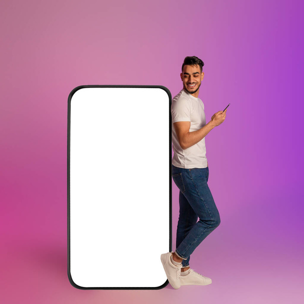 Happy young Arab man using mobile device while leaning on big smartphone with empty screen in neon light. Mockup - Photo, Image