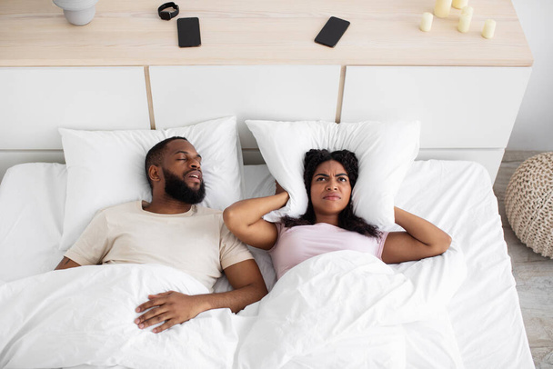 Upset despair millennial black woman covers ears with pillow, suffers from snoring of husband on bed - Foto, Bild