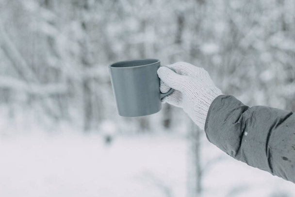 A hand in a white knitted woolen mitt and a gray jacket holds a gray mug against the background of a beautiful snow-covered forest. - Foto, Bild
