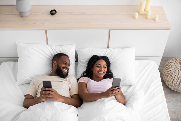 Glad smiling young black female and male, lying on bed, typing on phones, chatting, playing online game - Zdjęcie, obraz