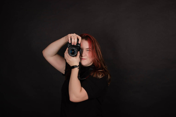 pelirroja con una cámara profesional posando sobre fondo negro. mujer fotógrafa.  - Foto, Imagen