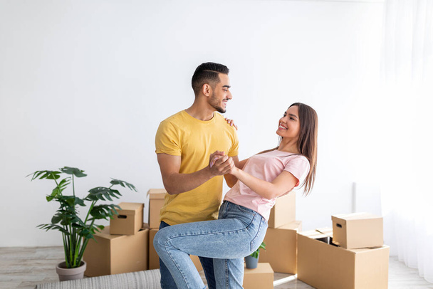 Feliz millennial pareja diversa bailando en su nuevo hogar entre cajas de cartón en el día de la reubicación, espacio de copia - Foto, imagen