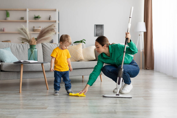 Little kid boy watching her mother mopping the floor after him. Woman wiping the floor with rag next to her baby - 写真・画像