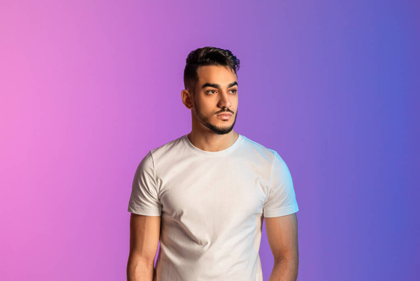 Portrait of serious young Arab man in white t-shirt posing and looking away in neon light - Fotografie, Obrázek