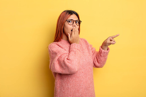 young hispanic woman feeling happy, shocked and surprised, covering mouth with hand and pointing to lateral copy space - 写真・画像