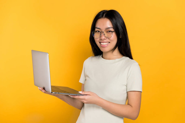 Smart belle fille brune chinoise aux cheveux longs, avec des lunettes, étudiante ou pigiste, tenant un ordinateur portable ouvert à la main, debout sur un fond orange isolé, regardant la caméra souriante amicale - Photo, image