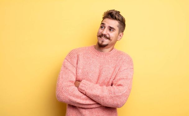 young handsome man feeling happy, proud and hopeful, wondering or thinking, looking up to copy space with crossed arms. strange moustache concept - Photo, Image