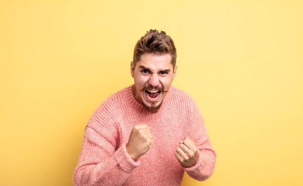 young handsome man shouting aggressively with annoyed, frustrated, angry look and tight fists, feeling furious. strange moustache concept - Photo, Image