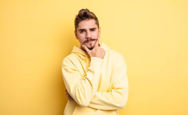 young handsome man looking serious, thoughtful and distrustful, with one arm crossed and hand on chin, weighting options. strange moustache concept - Photo, image
