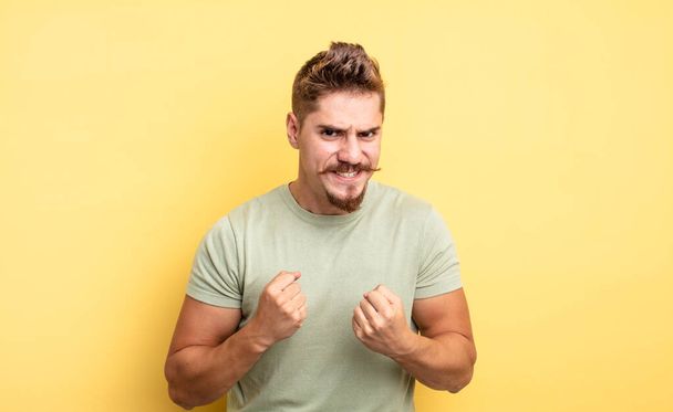young handsome man shouting aggressively with annoyed, frustrated, angry look and tight fists, feeling furious. strange moustache concept - Фото, изображение