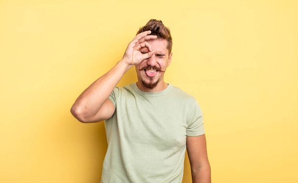 young handsome man smiling happily with funny face, joking and looking through peephole, spying on secrets. strange moustache concept - Photo, image