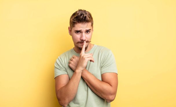 young handsome man looking serious and cross with finger pressed to lips demanding silence or quiet, keeping a secret. strange moustache concept - Foto, afbeelding