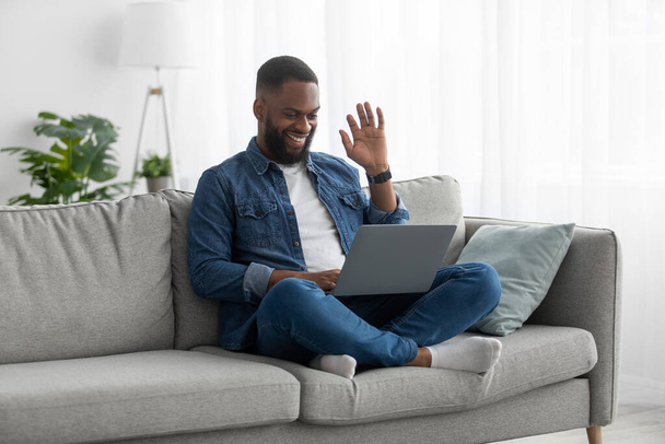 Happy attractive young african american male with beard sits on couch, greets, waves his hand to pc webcam - Фото, зображення