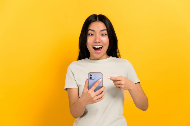 Joyeux, surpris étonné asiatique aux cheveux longs fille brune, dans un t-shirt décontracté, tient un smartphone dans sa main pointe du doigt à elle, regarde étonné dans la caméra, se tient sur un fond orange isolé - Photo, image