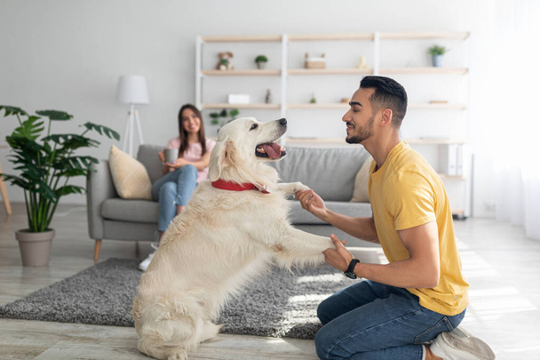 幸せな若いアラブ人の男は犬と遊んで、家で床で楽しんで、彼のガールフレンドはバックグラウンドでコーヒーを飲む - 写真・画像