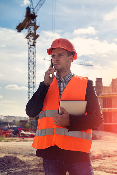 een jonge werknemer in uniform en helm op een nieuw gebouw belt de baas om de details van de bouw te verduidelijken. Bouwconcept - Foto, afbeelding