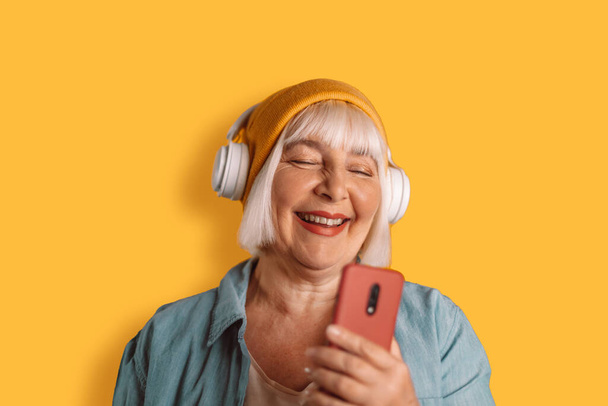 Happy 50s nainen kuulokkeet kuunnella musiikkia laulaa laulun ennätys ääni matkapuhelin sanelee keltainen väri tausta studio muotokuva. - Valokuva, kuva