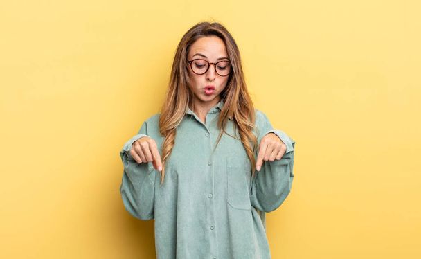 pretty caucasian woman with open mouth pointing downwards with both hands, looking shocked, amazed and surprised - 写真・画像
