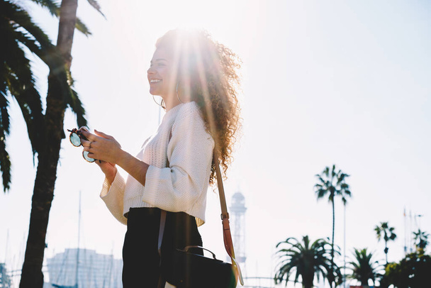 Backlit of pretty female user holding modern smartphone gadget in hands enjoying summer vacations, side view of happy curly blogger with cellphone for online networking smiling at sunny street - Photo, Image
