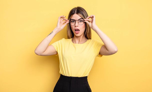pretty caucasian woman feeling shocked, amazed and surprised, holding glasses with astonished, disbelieving look - Foto, afbeelding