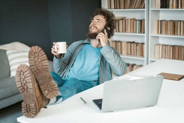 Positive Businessman in Casual Clothes has a Easy Phone Conversation with a Cup of Coffee in his Hand and Feet on the Table. Cute Caucasian Guy Talking on Smartphone in the Office. Close-up - 写真・画像