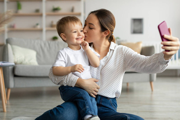 Madre cariñosa tomando selfie con su hijo pequeño y besando a un niño en la mejilla, mamá capturando una foto en un teléfono inteligente - Foto, Imagen