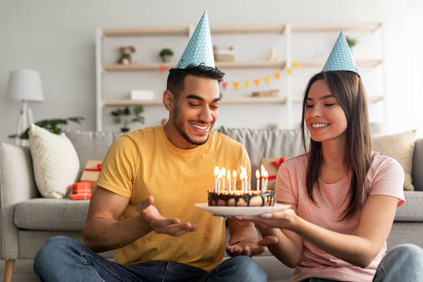 Cariñosa pareja multirracial milenaria en sombreros de fiesta sosteniendo pastel de cumpleaños con velas encendidas en la sala de estar - Foto, Imagen