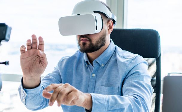 Bearded focused male entrepreneur using virtual reality goggles while sitting on office chair at table in modern workplace during work - Photo, image