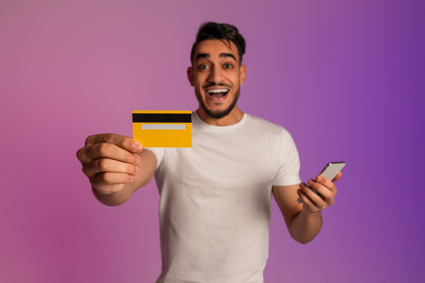 Online shopping app. Joyful millennial Arab man holding cellphone, showing credit card in neon light, selective focus - Fotoğraf, Görsel