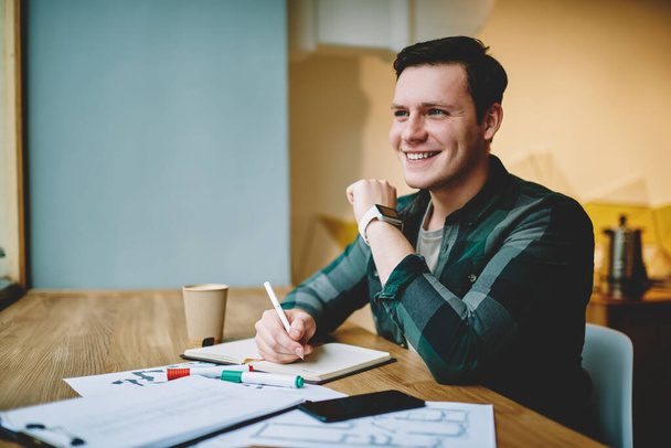 Young happy male freelancer in casual outfit taking notes in planner sitting at table with drawings smartphone and paper cup in cafe - Foto, Imagem