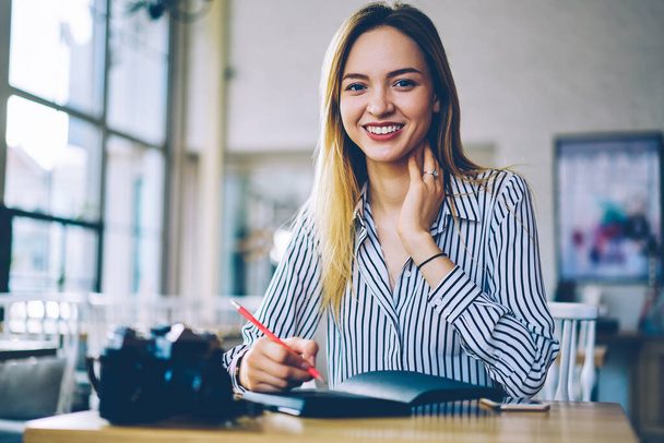 Portrait of cheerful Caucasian woman smiling at camera while spending leisure time for organisation planning in coffee shop, happy female student with education textbook posing at cafeteria table - Φωτογραφία, εικόνα