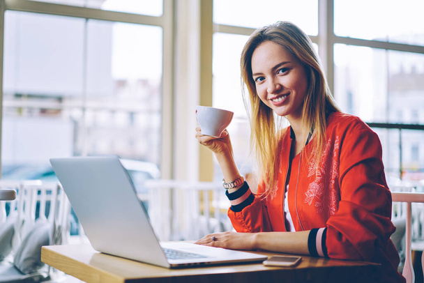 カフェで遠隔作業のための現代のラップトップコンピュータを使用してカメラで笑顔おいしいコーヒーのカップと陽気な女性のコピーライターの肖像,幸せな白人学生電子書籍で学ぶ - 写真・画像