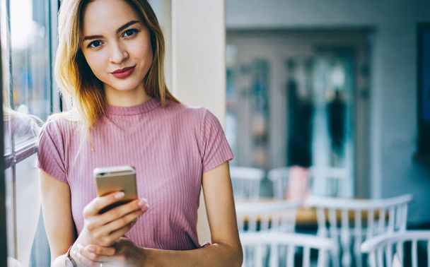 Half length portrait of attractive young woman blogger with modern cellphone technology in hands looking at camera during leisure pastime, beautiful female millennial with mobile phone posing indoors - Photo, Image