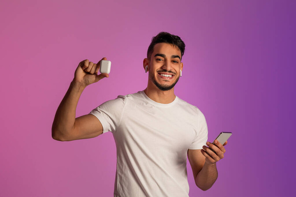 Smiling Arab guy with smartphone advertising wireless earphopnes, listening to music via new mobile app in neon light - Fotografie, Obrázek