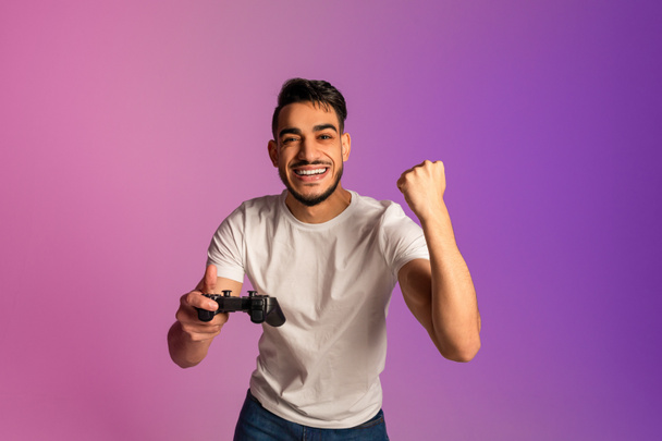 Excited millennial Arab man with joystick celebrating video game win, gesturing YES in neon light - Valokuva, kuva