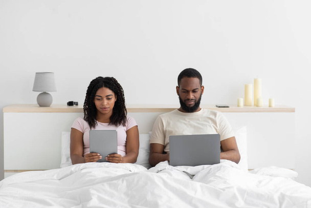 Зайнята молода чорна жінка з планшетом ігнорує чоловіка з комп'ютером, сидить на ліжку в інтер'єрі спальні ввечері
 - Фото, зображення