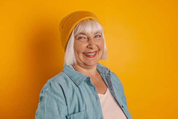 Photo of optimistic older blond lady in a yellow hat and stylish casual clothes smiling at the camera isolated over color background, People lifestyle concept - Photo, Image