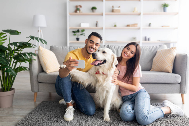 Young international couple taking mobile selfie with pet dog on smartphone at home, copy space - Foto, Bild