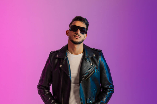 Portrait of cool serious young Arab man in sunglasses wearing black leather jacket, looking at camera in neon light - Foto, afbeelding