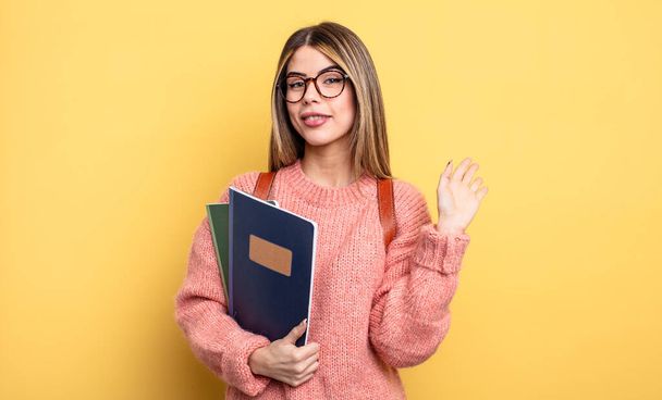 pretty student woman smiling happily, waving hand, welcoming and greeting you. books and backpack - 写真・画像