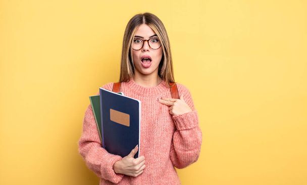 pretty student woman looking shocked and surprised with mouth wide open, pointing to self. books and backpack - Zdjęcie, obraz