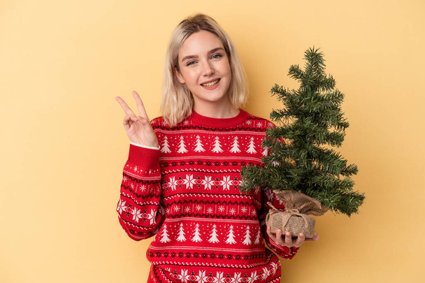 Joven mujer caucásica sosteniendo un pequeño árbol de navidad aislado sobre fondo amarillo alegre y despreocupado mostrando un símbolo de paz con los dedos. - Foto, imagen