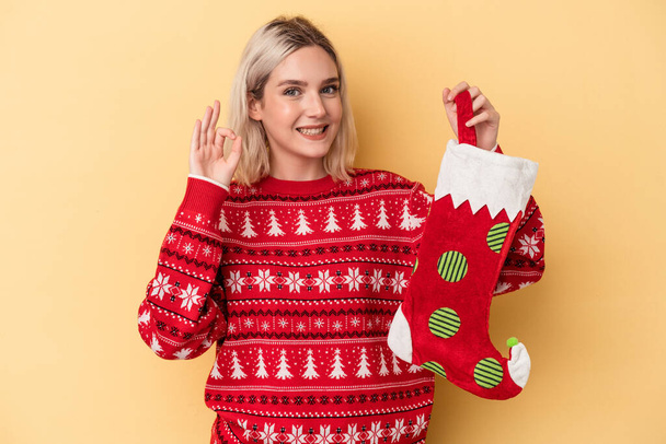 Young caucasian woman holding a elf sock isolated on yellow background cheerful and confident showing ok gesture. - Photo, Image