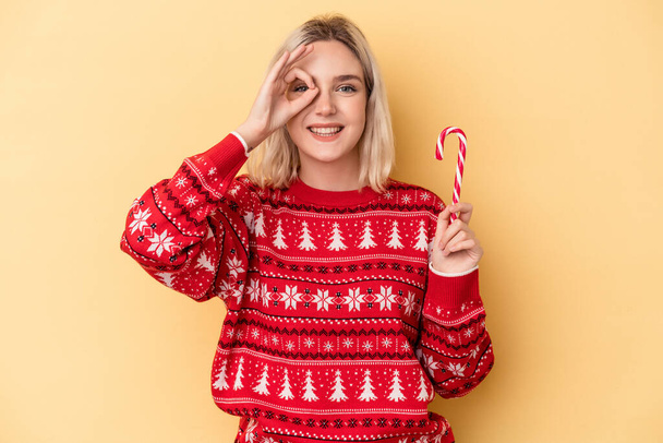 Young caucasian woman holding a christmas stick isolated on yellow background excited keeping ok gesture on eye. - Photo, Image