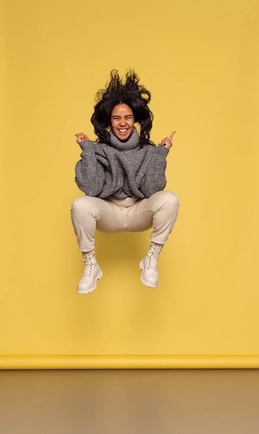 Full-length portrait of young happy dark skinned girl in warm knitted sweater jumping isolated on yellow background. - Photo, image