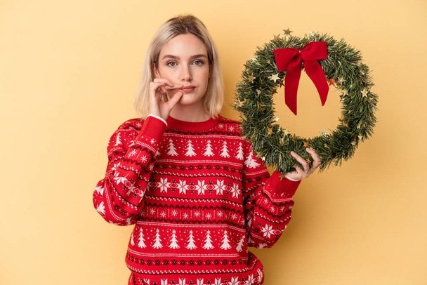 Joven mujer caucásica sosteniendo una corona de Navidad aislada sobre fondo amarillo con los dedos en los labios manteniendo un secreto. - Foto, imagen