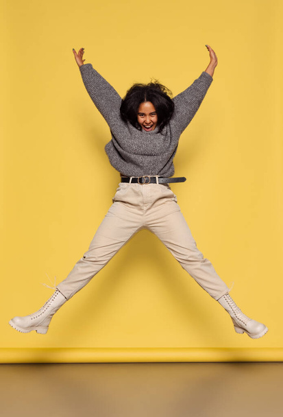 Full-length portrait of young happy dark skinned girl in warm knitted sweater jumping isolated on yellow background. - Φωτογραφία, εικόνα